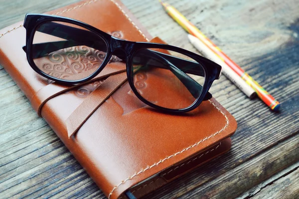 Un viejo cuaderno en funda de cuero con lápices y vasos sobre mesa de madera — Foto de Stock