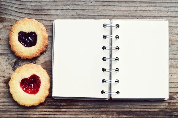 Biscoitos caseiros com geléia em forma de coração e livro de receitas espiral retro vazio na mesa de madeira — Fotografia de Stock