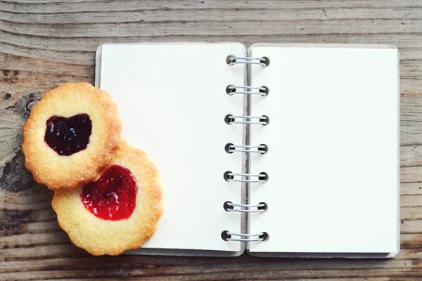 Galletas caseras con mermelada en forma de corazón y libro de recetas en espiral retro vacío en mesa de madera — Foto de Stock