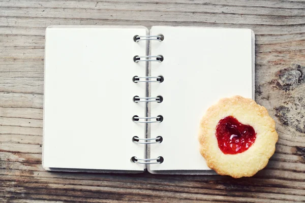 Homemade cookies with heart shaped jam and empty retro spiral recipe book on wooden table — Stock Photo, Image