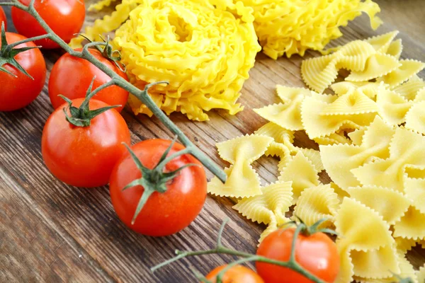 Bunch of small red cherry tomatoes with raw pasta Reginette (Mafaldine) and butterfly shaped pasta farfalle on wooden table