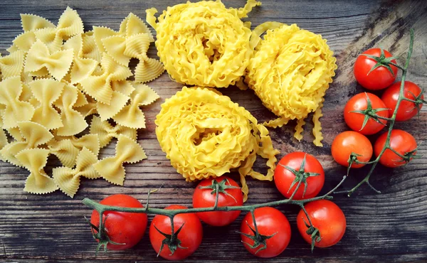 Bunch of small red cherry tomatoes with raw pasta Reginette (Mafaldine) and butterfly shaped pasta farfalle on wooden table