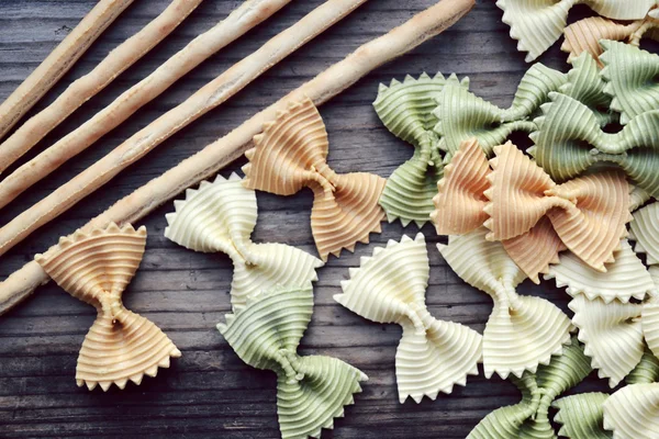 Borboleta não cozida em forma de farfalle de macarrão colorido com breadsticks na mesa de madeira — Fotografia de Stock