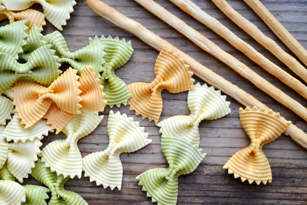 Pâtes alimentaires colorées en forme de papillon non cuites farfalle avec des bâtonnets de pain sur une table en bois — Photo