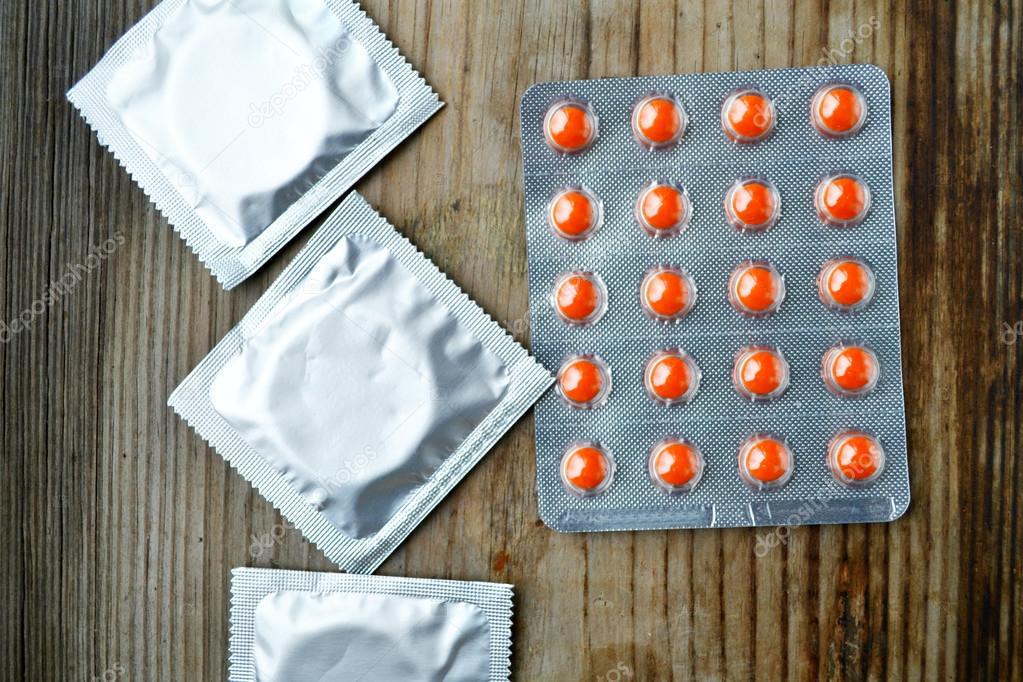 Three condoms and orange birth control pills on wooden table