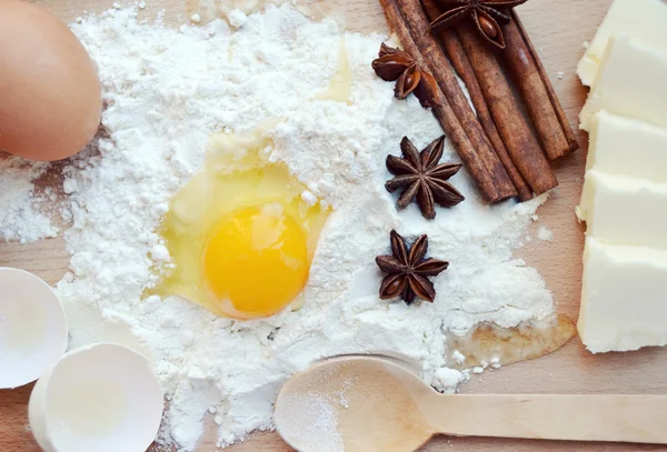 White flour with eggs, butter, cinnamon sticks, spicy anise stars and wooden spoon on a cooking board — Stock Photo, Image