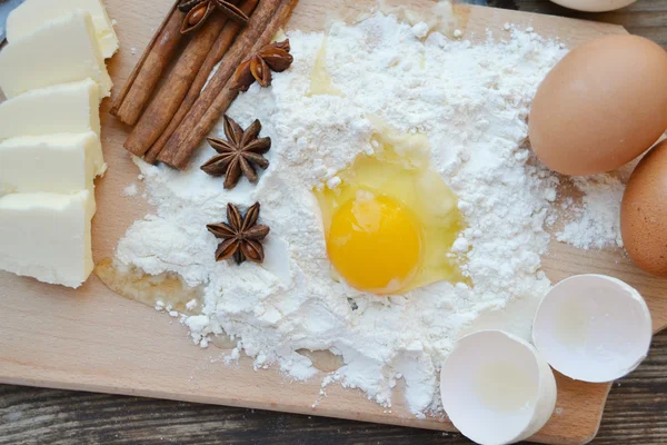 White flour with eggs, butter, cinnamon sticks, spicy anise stars and wooden spoon on a cooking board — Stock Photo, Image