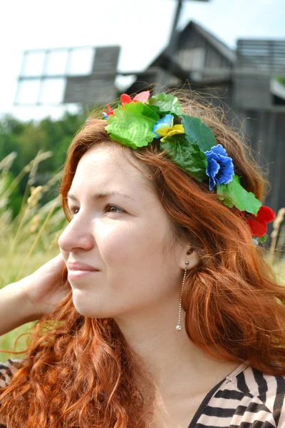 Vakker ung rødhåret med blomster i håret, kvinne som smiler lykkelig med en mølle i bakgrunnen. – stockfoto