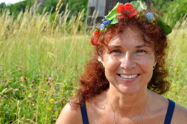 Mujer pelirroja hermosa feliz con flores en el pelo en sus cuarentas oliendo al aire libre — Foto de Stock