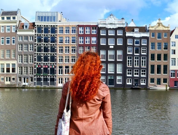 Mujer pelirroja en chaqueta de cuero de pie con su espalda a la cámara y mirando a los edificios tradicionales de la ciudad de Amsterdam cerca del canal y los barcos — Foto de Stock