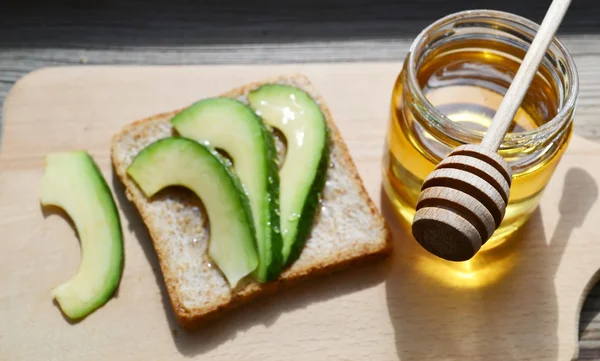 Golden honey with honeystick and a healthy sandwich with green avocado on wooden board — Stock Photo, Image