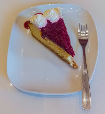 Krem şanti ile süslenmiş kırmızı çilekli kek. Kare beyaz bir porselen tabakta çatalın yanında servis ediliyor..