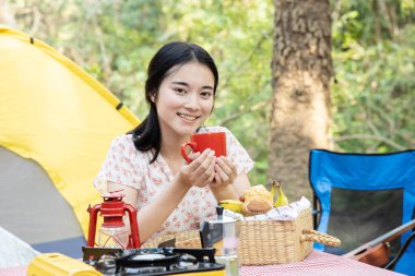 An Asian woman drinking coffee in front of a tent, enjoying a camping