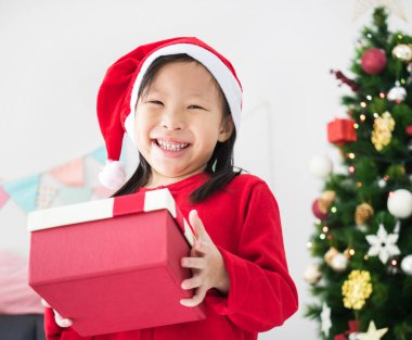 Mutlu Asyalı küçük kız elinde hediye kutusuyla Noel ağacının önünde duruyor