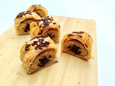 Delicious golden brown Indonesian bolen pisang, a flaky puff pastry filled with sweet banana, topped with chocolate sprinkles, presented on a rustic wooden board.