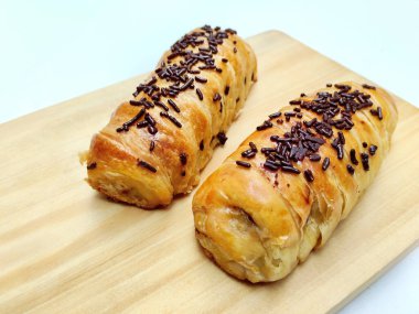 Delicious golden brown Indonesian bolen pisang, a flaky puff pastry filled with sweet banana, topped with chocolate sprinkles, presented on a rustic wooden board.