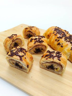Delicious golden brown Indonesian bolen pisang, a flaky puff pastry filled with sweet banana, topped with chocolate sprinkles, presented on a rustic wooden board.