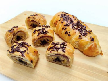 Delicious golden brown Indonesian bolen pisang, a flaky puff pastry filled with sweet banana, topped with chocolate sprinkles, presented on a rustic wooden board.