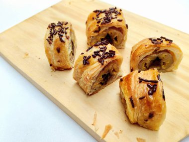 Delicious golden brown Indonesian bolen pisang, a flaky puff pastry filled with sweet banana, topped with chocolate sprinkles, presented on a rustic wooden board.