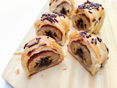 Delicious golden brown Indonesian bolen pisang, a flaky puff pastry filled with sweet banana, topped with chocolate sprinkles, presented on a rustic wooden board.