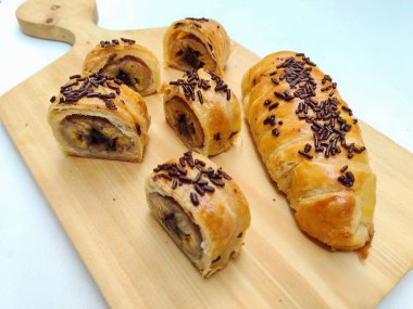 Delicious golden brown Indonesian bolen pisang, a flaky puff pastry filled with sweet banana, topped with chocolate sprinkles, presented on a rustic wooden board.