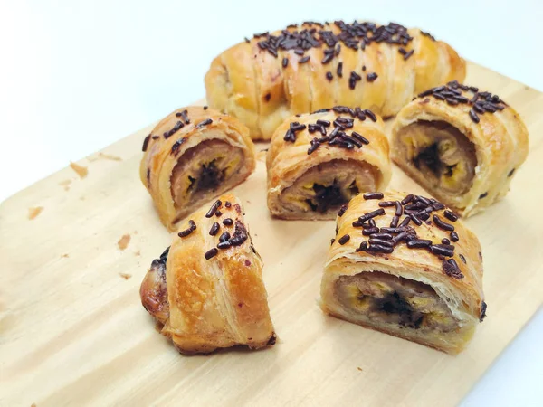 Delicious golden brown Indonesian bolen pisang, a flaky puff pastry filled with sweet banana, topped with chocolate sprinkles, presented on a rustic wooden board.