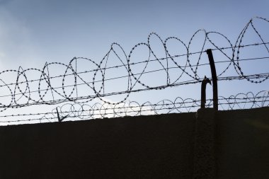 Hapishane duvarlarının etrafındaki dikenli tel örgü. Arka planda mavi gökyüzü.