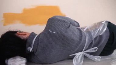 Uzanan kız, duvarı fırçayla turuncuya boyuyor.