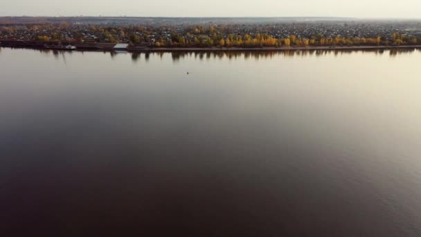 无人机飞过一条宽阔的河流 岸边映衬着秋天的树木 这个村子在远处看得见 — 图库视频影像