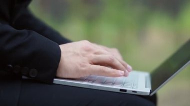 Takım elbiseli bir adam ormanda otururken dizüstü bilgisayarına mesaj basıyor. Yakın plan.