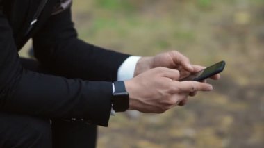 Takım elbiseli bir adam parktaki bankta otururken telefonunun ekranına bakar. Yakın plan.