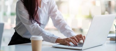 Bronzlaşmış bir kadının ellerini bilgisayar klavyesine yakın tut. O daktilo ediyor. Üzerinde mavi grafikler ve bir fincan kahve olan bir kağıt parçası var.