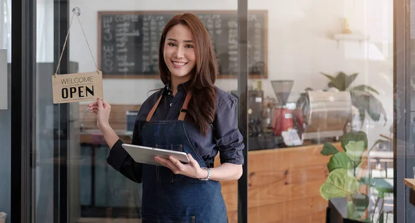 Portrait of a smiling Asian entrepreneur standing behind her cafe counter with open sign boar