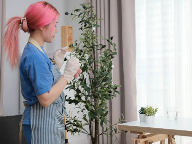 Mavi önlüklü bir kadın üstünde laptop olan bir masayı temizliyor. Elinde bir şişe temizleyici de var.