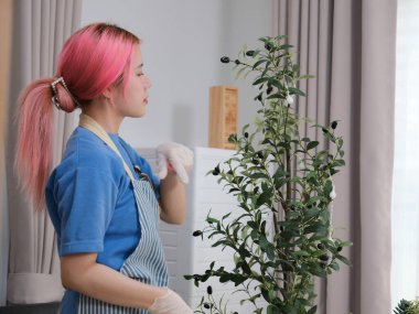 Mavi önlüklü bir kadın üstünde laptop olan bir masayı temizliyor. Elinde bir şişe temizleyici de var.