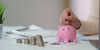 Bir kadın, modern bir çalışma alanındaki mali planlama ve tasarrufları simgeleyen pembe bir domuz kumbarasına para yatırıyor..