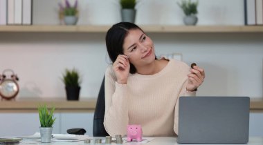 Genç bir kadın elinde bozuk para tutarken gülümsüyor. Parlak bir ofisteki mali yolculuğunu ve tasarruf stratejisini yansıtıyor..
