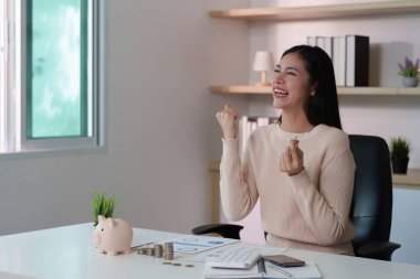 Paralar ve kumbarayla çevrili, finansal başarılarından dolayı heyecanlanan neşeli bir kadın..