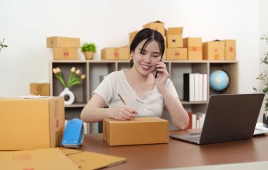Genç bir kadın, teslimat için paketleri hazırlarken telefonda konuşuyor. Küçük işletmelerinde birden fazla görevi olduğunu gösteriyor..
