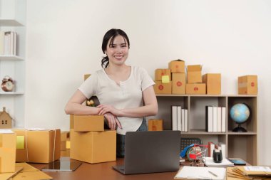 Genç bir kadın, modern e-ticaret ortamını temsil eden paketlerle çevrili olarak, güvenli bir şekilde ev ofisinde duruyor..