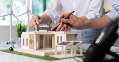 İki kişi bir ev modeli üzerinde çalışıyor. Biri bir kalem tutuyor ve diğeri de evi ölçüyor. Sahne odaklı ve ciddi.