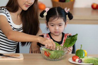 Bir kadın küçük bir kıza salatadan sebze seçmesinde yardım ediyor. Kız üzgün bir ifadeyle salataya bakıyor.