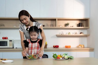 Bir kadın ve küçük bir kız mutfakta yemek hazırlıyorlar. Kadın, kızın sebzeleri kaseye koymasına yardım ediyor. Sahne sıcak ve besleyici, çünkü kadın kıza yemek pişirmeyi öğretiyor.