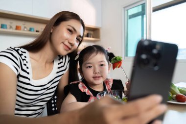 Bir kadın ve genç bir kız önlerinde cep telefonuyla bir masada oturuyorlar. Kadın, elinde telefon olan kızın fotoğrafını çekiyor. Sahne mutlu ve sevgi dolu.