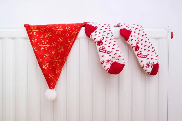 Біло-червоні різдвяні шкарпетки та капелюх Санти, що звисають на теплому центральному тепловому радіаторі, зимовий домашній настрій Стокове Фото
