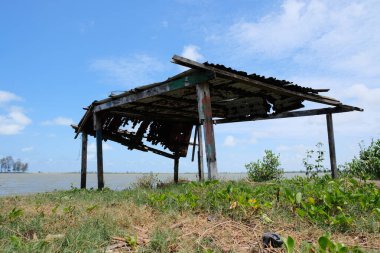 Kırık paslı çatısı olan harap bir ahşap kulübe, Malezya 'nın Kelantan kentindeki açık mavi gökyüzünün altında nehir kenarında duruyor..
