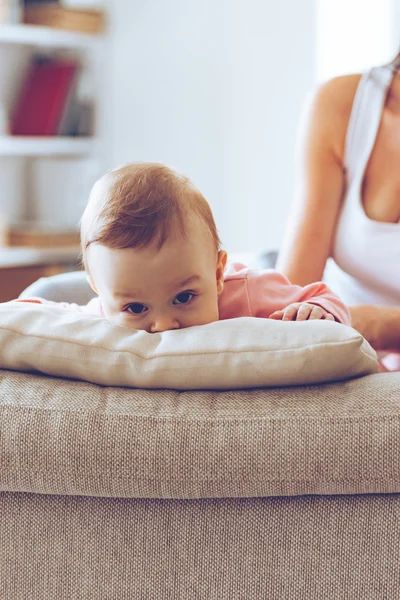 Her look can melt any heart! Close-up part of little baby girl looking at camera and leaning on pillow while her mother sitting on the couch at background — Stock Photo, Image