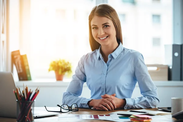 Sonrisa dentada de la experiencia real. Joven alegre hermosa mujer mirando a la cámara con sonrisa mientras está sentada en su lugar de trabajo — Foto de Stock