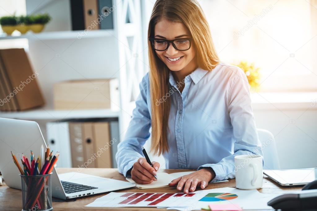 Expert in her business. Cheerful young beautiful woman making some notes in notebook and looking at charts with smile while sitting at her working place