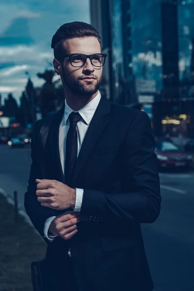 Un hombre de negocios moderno. Joven confiado en traje completo ajustando su manga y mirando hacia otro lado mientras está parado al aire libre con paisaje urbano en el fondo — Foto de Stock